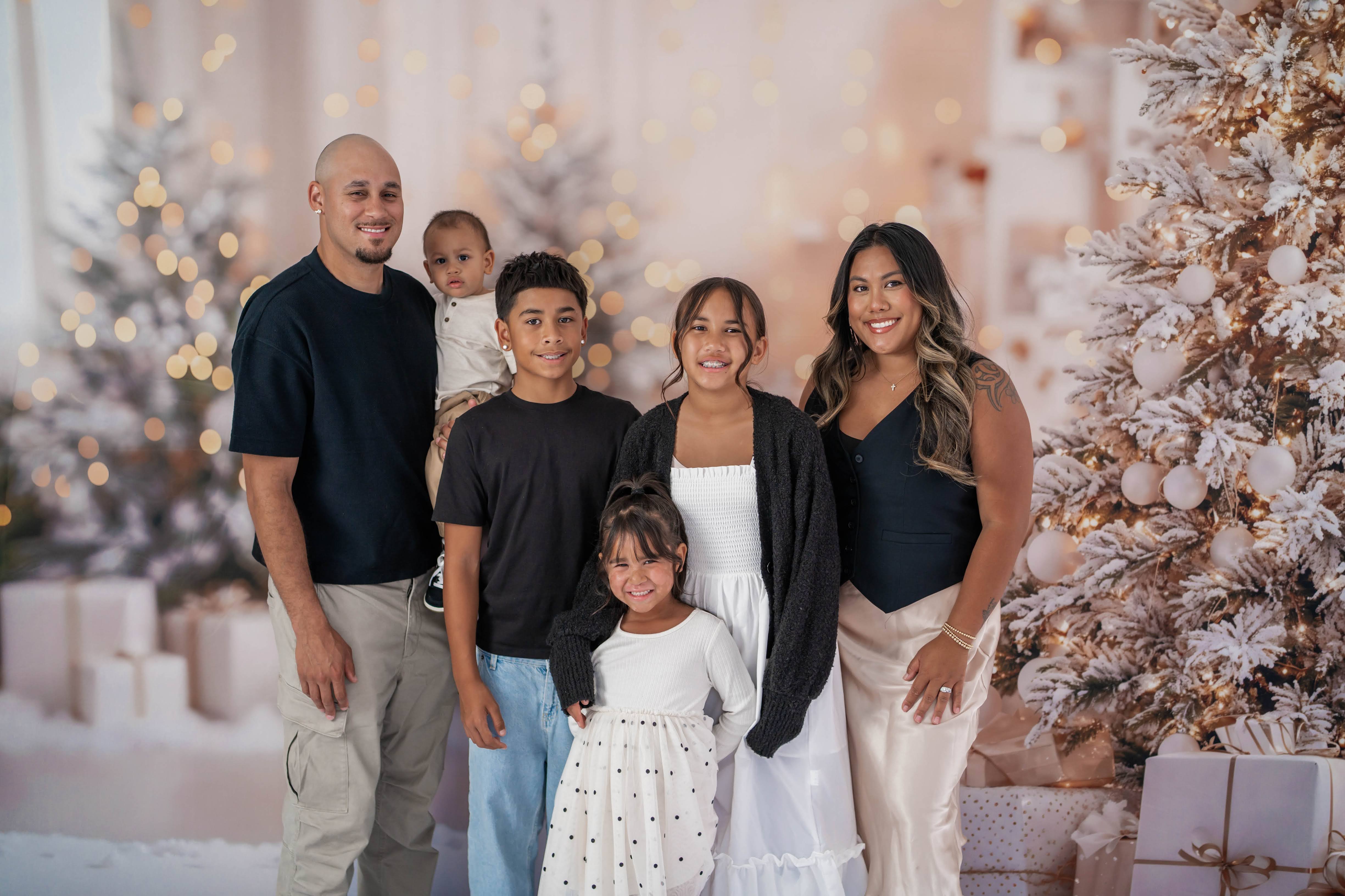 Family of six posing beside flocked Christmas trees, warm lights, and wrapped gifts.