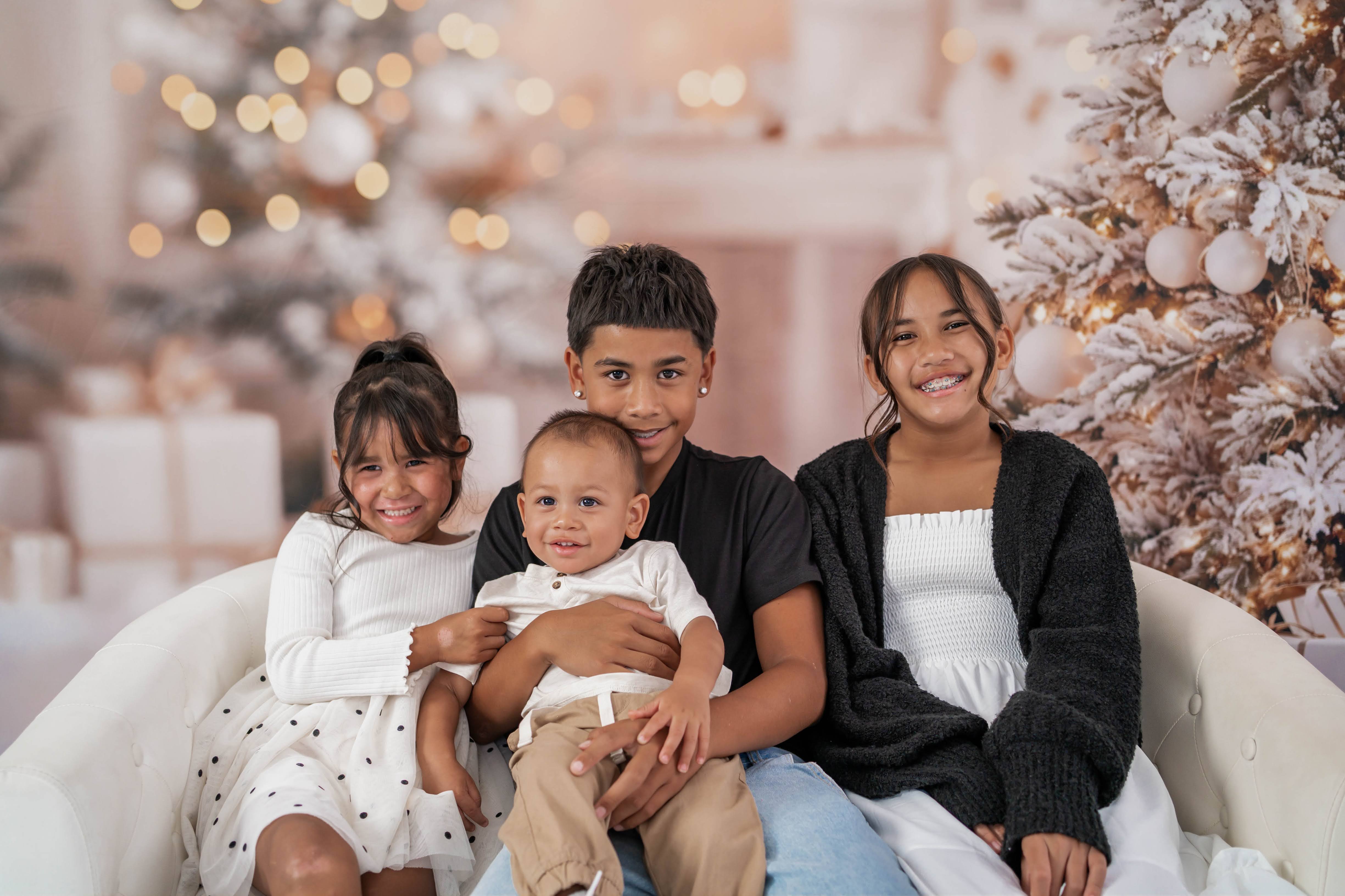 Four siblings sitting close together on a cream sofa beside a flocked Christmas tree.