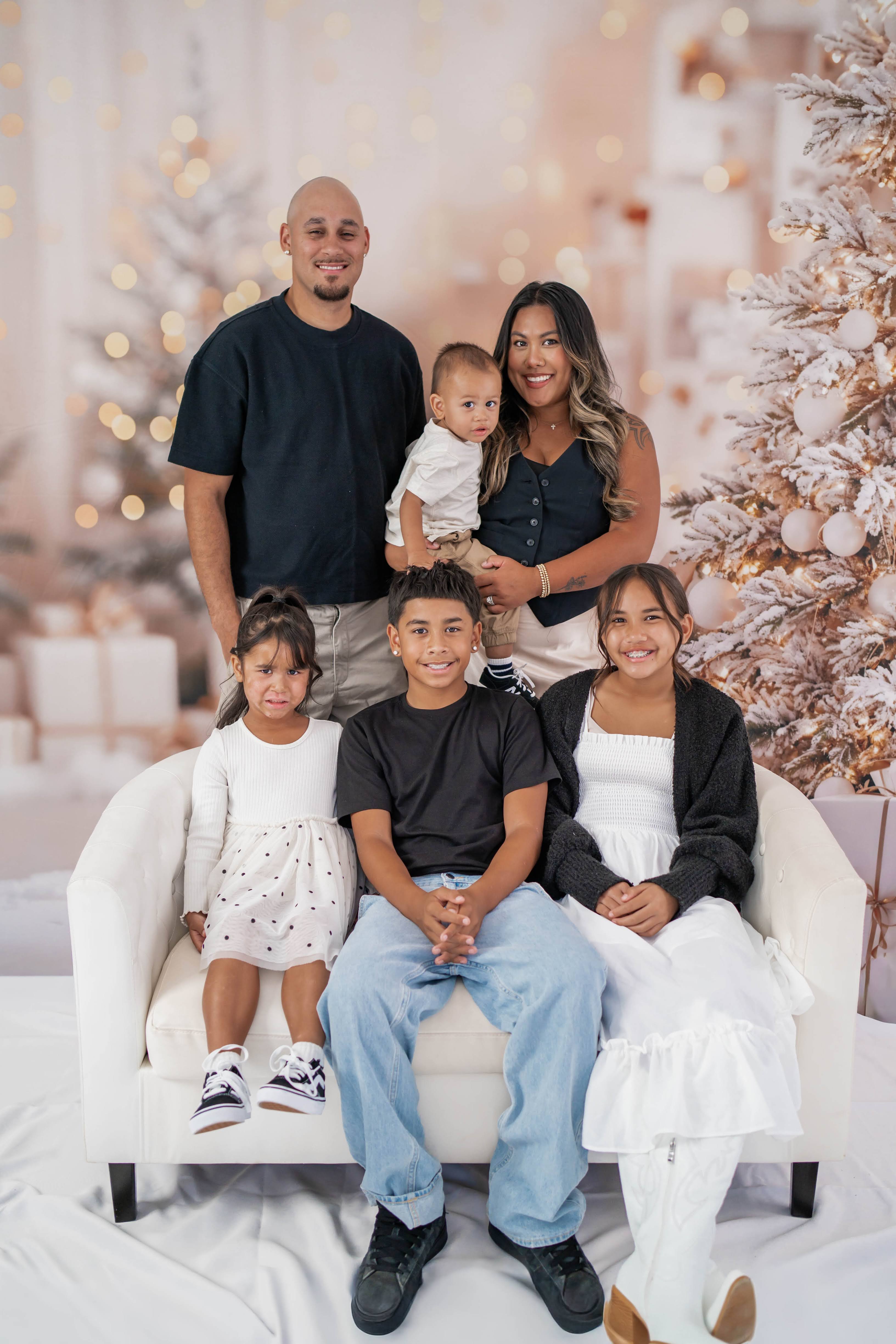 Family of six gathered on and around a cream sofa in front of flocked Christmas trees and warm lights.