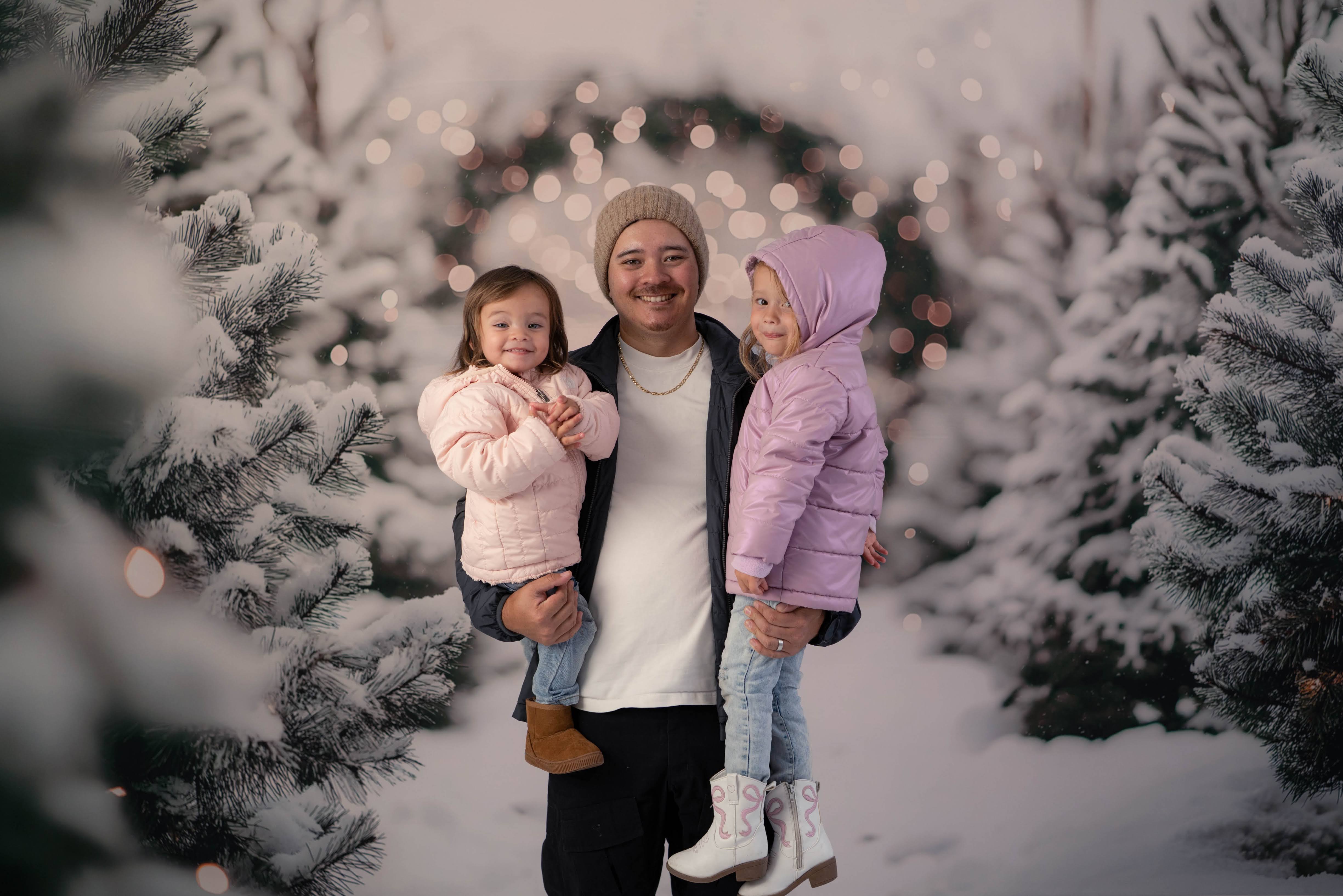 Man holding two young children between snow-covered Christmas trees and warm lights.