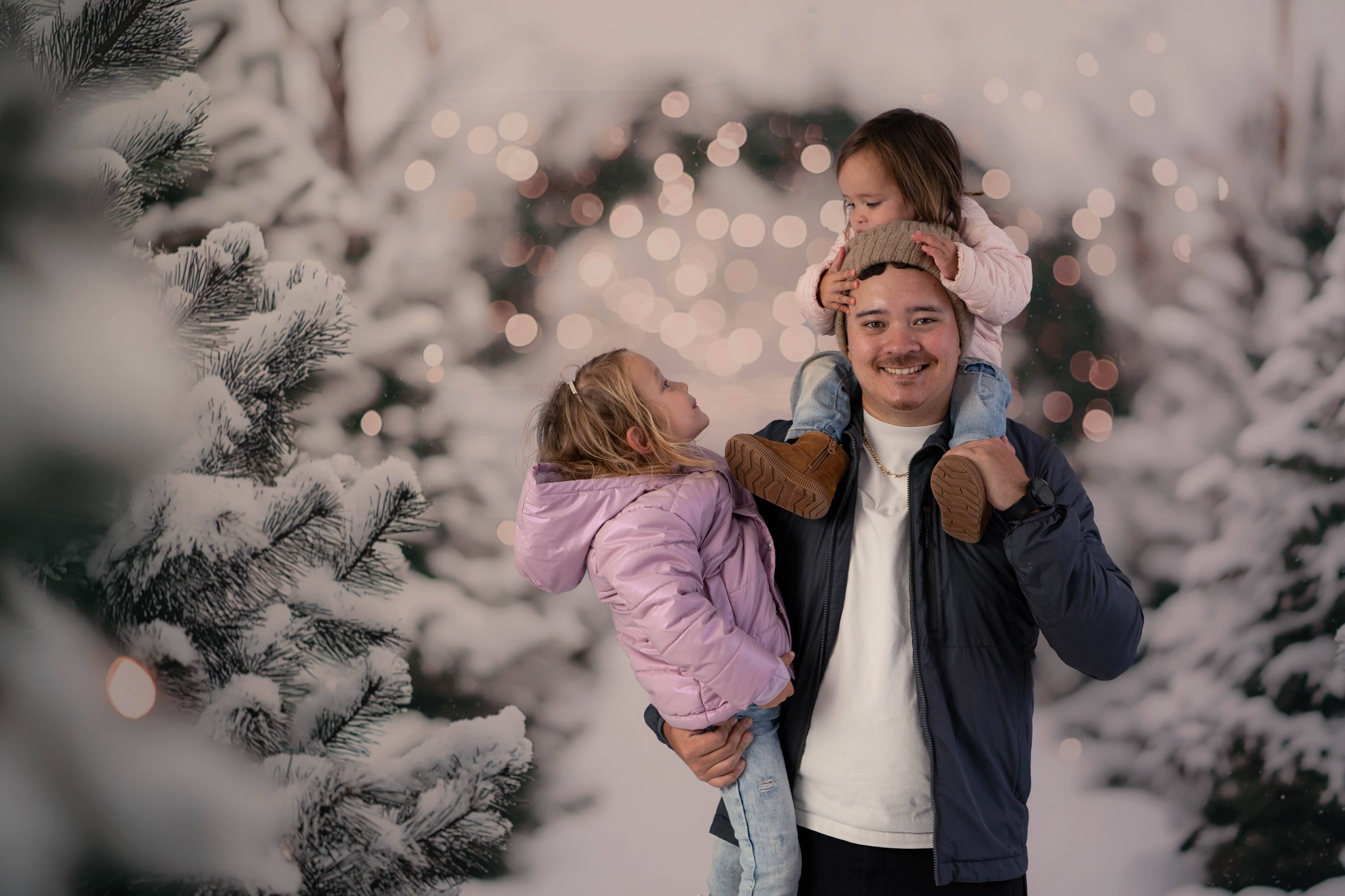 Dad carrying one child on his shoulders and another in his arms between snow-covered evergreens.