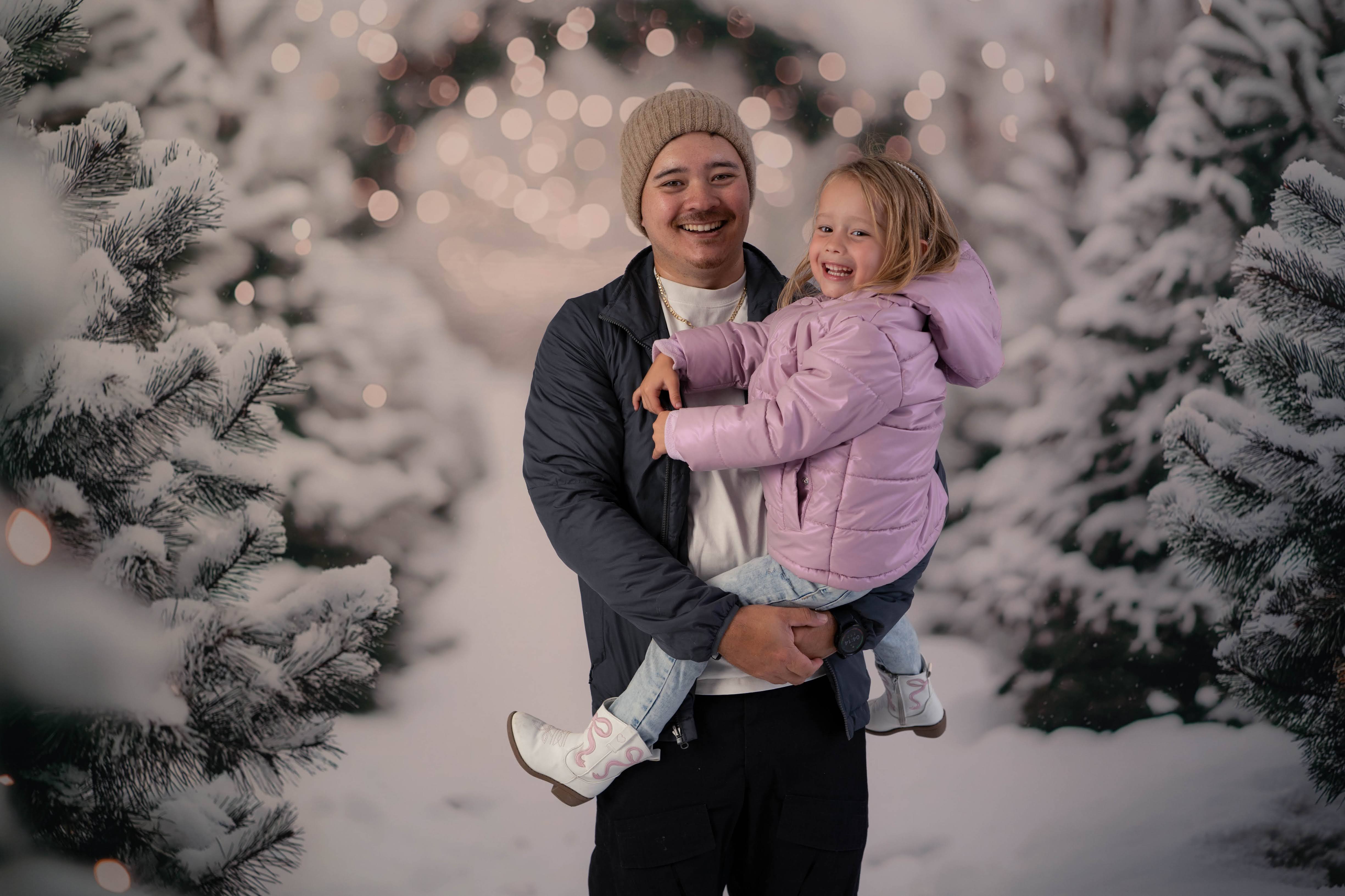Smiling dad holding his daughter between snow-covered Christmas trees and soft holiday lights.