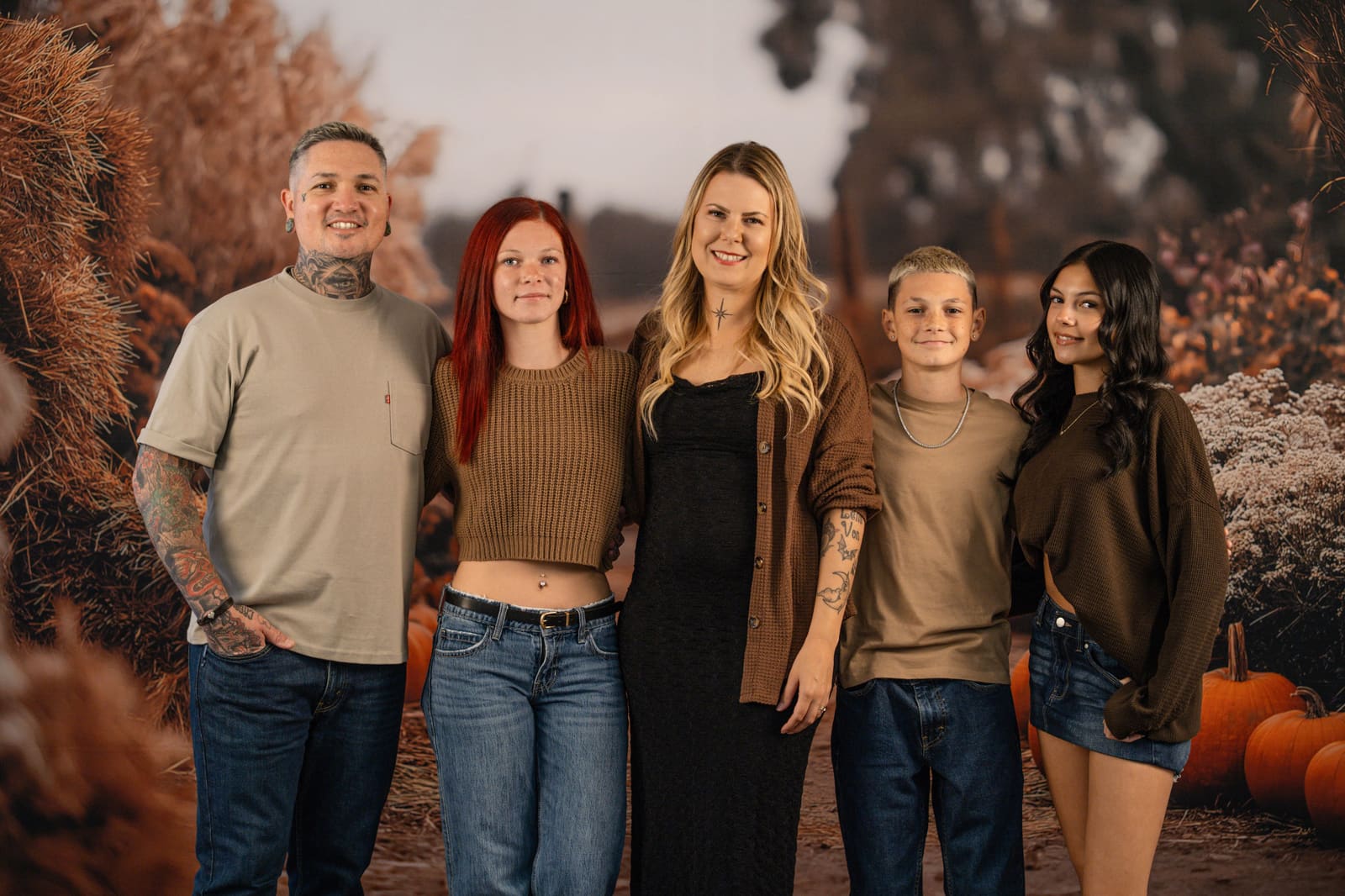 Family of five standing together in the harvest field set with pumpkins and warm fall grasses.