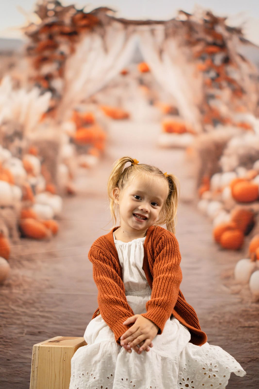 Preschooler in a rust cardigan sitting on a crate under the pumpkin patch arch.