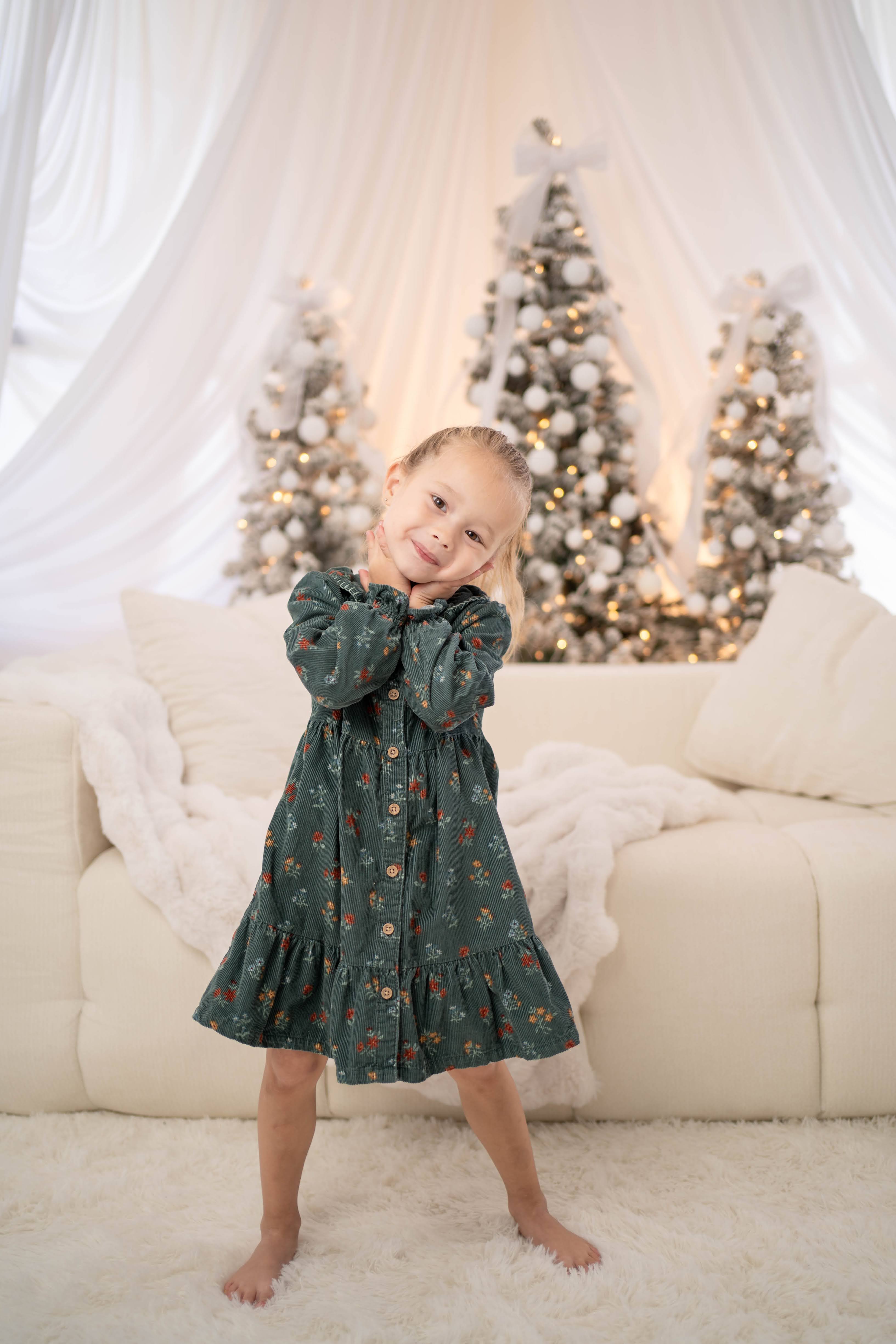 A young child in a dark green floral dress poses barefoot on a white rug in front of softly lit Christmas trees decorated with white ornaments, framed by white drapery and a cream sofa.