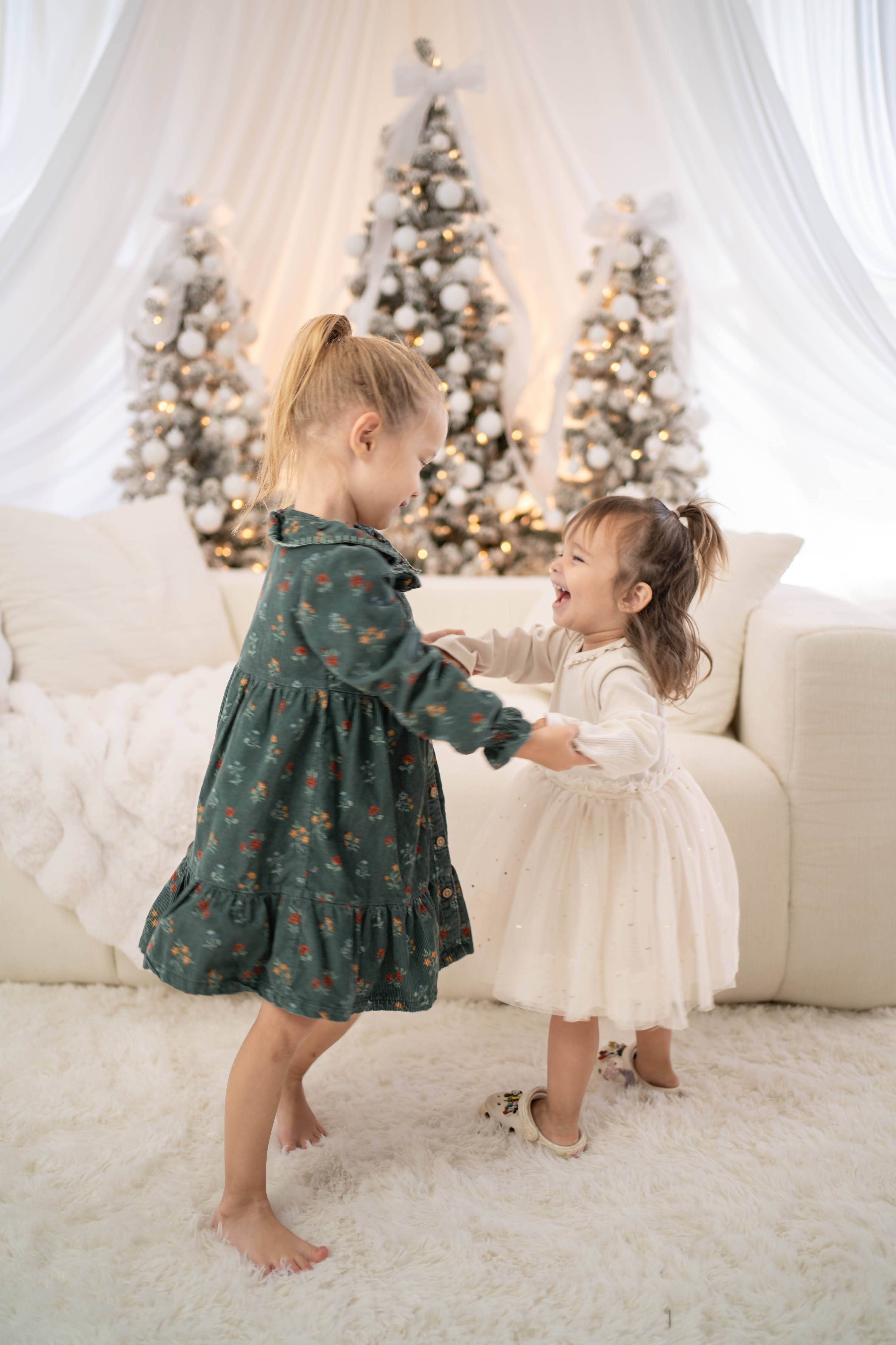 A child poses in a cozy all-white Christmas setting with warm twinkle lights and soft cream furniture.
